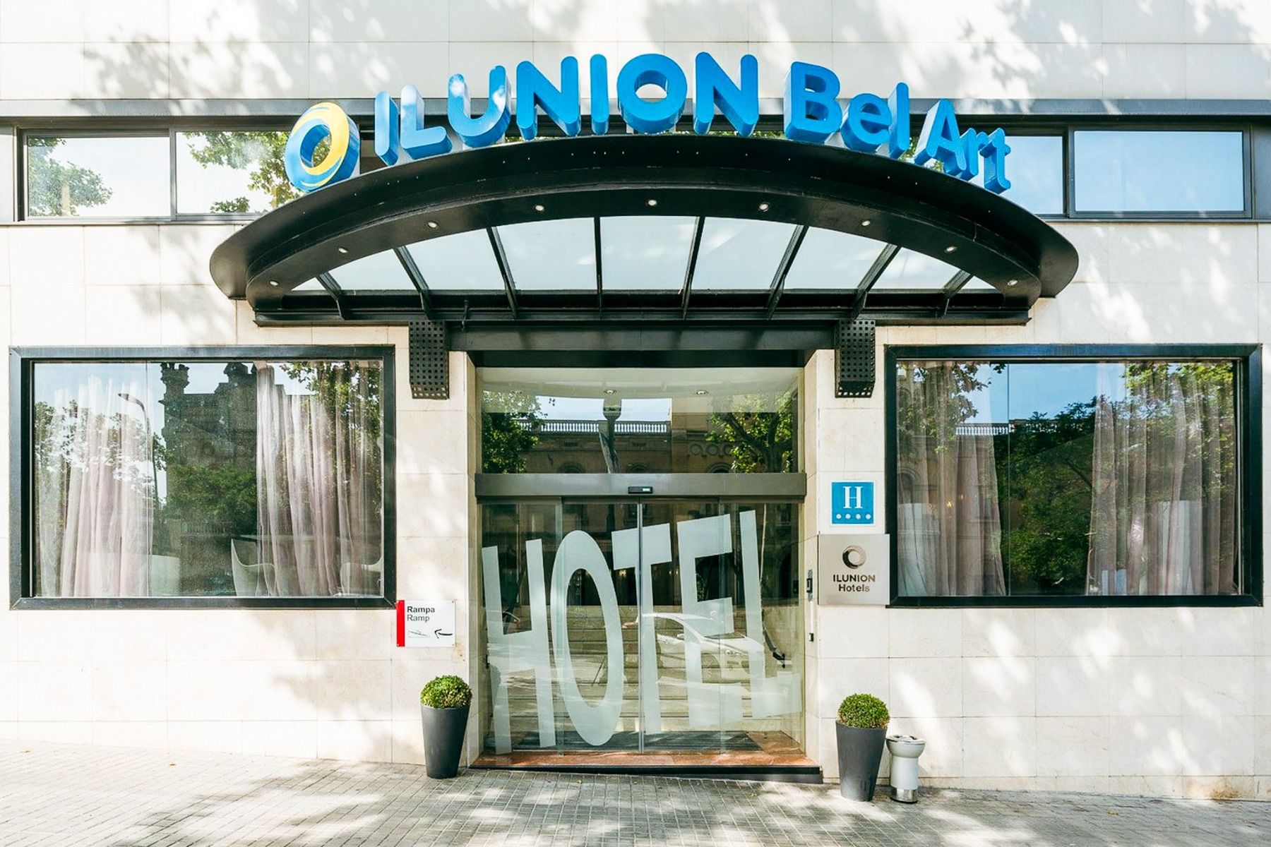 Inngangen til Hotel ILUNION Bel Art Barcelona har store glassdører, potteplanter og et skilt over den svarte markisen - perfekt for gjester som vil utforske Fußballkultur Barcelona eller besøke Trainingseinrichtungen i nærheten.