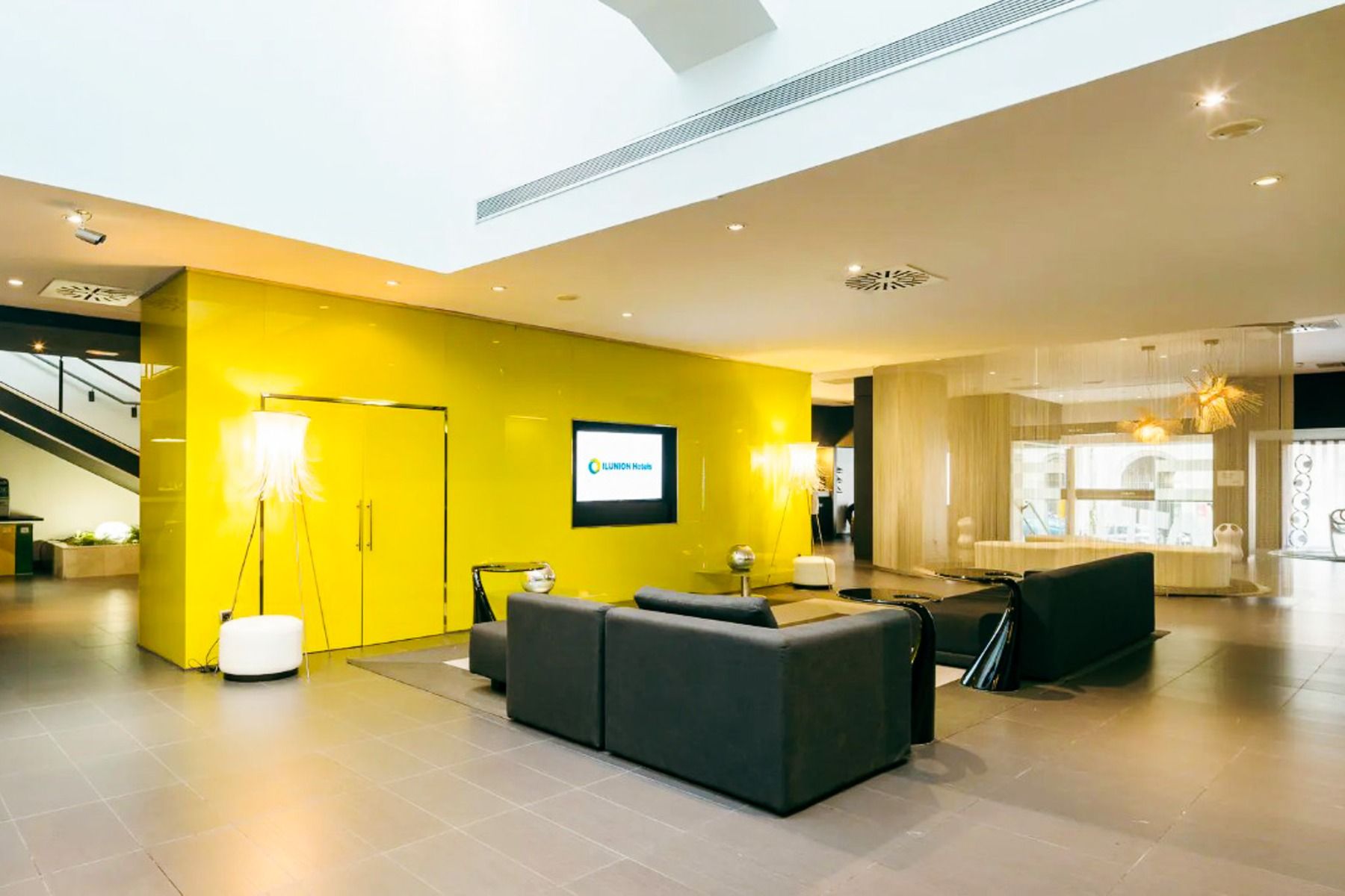 Lobbyen på Hotel ILUNION Bel Art Barcelona har svarte sofaer, et salongbord i glass, en iøynefallende gul vegg med en montert TV, myk belysning og moderne innredning som gjenspeiler Barcelonas pulserende fotballkultur. En trapp er også synlig.