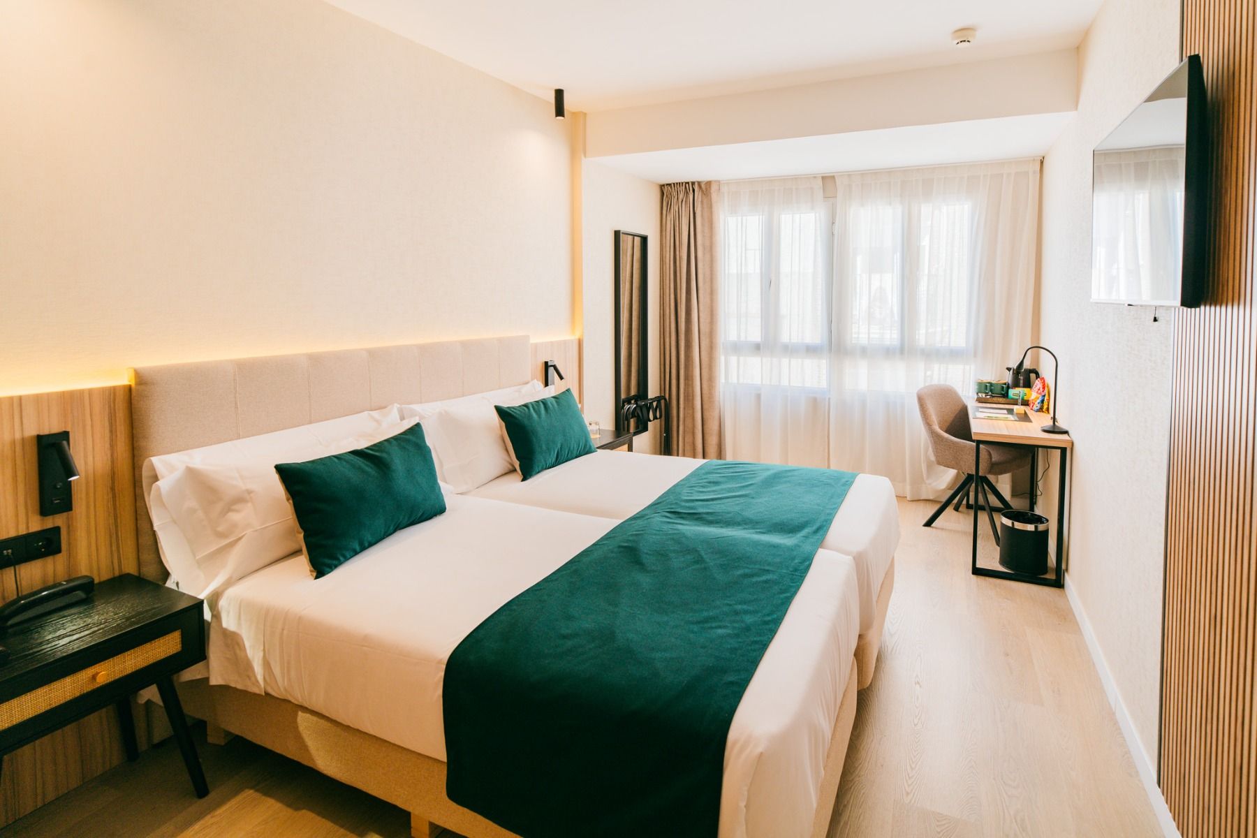 Ein Zimmer im Hotel Gaudea in Santa Pola bietet zwei Einzelbetten mit weißer Bettwäsche und grünen Akzenten, einen Schreibtisch am großen Fenster mit durchsichtigen Vorhängen und einen an der Wand befestigten Fernseher für Ihren Komfort.