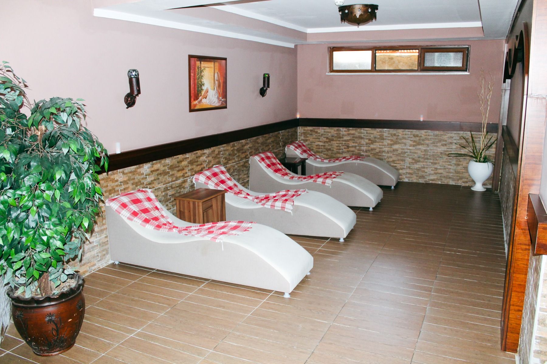 Der Wellnessraum des Linda Resorts ist mit fünf weißen Liegestühlen mit rot-weiß karierten Handtüchern, einer Topfpflanze, Wanddekoration, Fliesenboden und sanfter Beleuchtung ausgestattet.