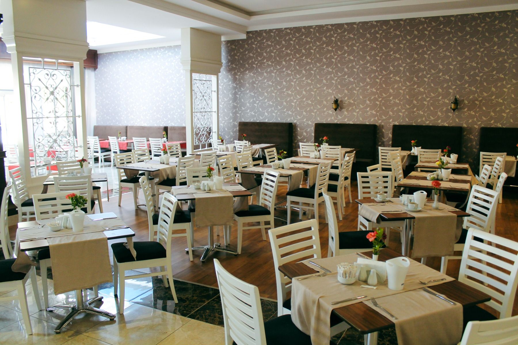 Das Linda Resort verfügt über einen modernen, hell beleuchteten Speiseraum mit weißen Holzstühlen, Tischen, die mit beigen Tischläufern geschmückt sind, Geschirr und kleinen Blumenvasen. Gemusterte Tapeten und große Fenster lassen den Raum in natürliches