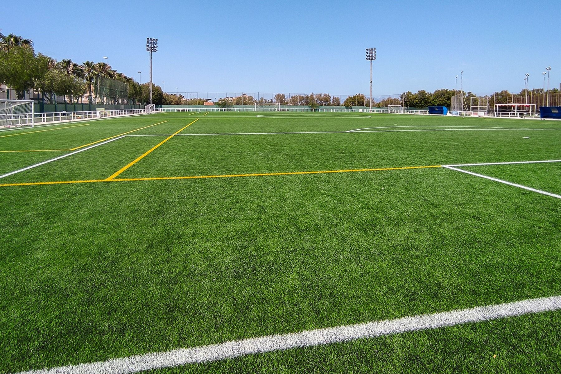 Ein großes, leeres Fußballfeld mit leuchtend grünem Kunstrasen und weißen und gelben Markierungen unter einem klaren blauen Himmel in der Nähe des Hotels Gaudea in Santa Pola. Im Hintergrund sind Flutlicht und Bäume neben dem 4-Sterne-Hotel zu sehen.