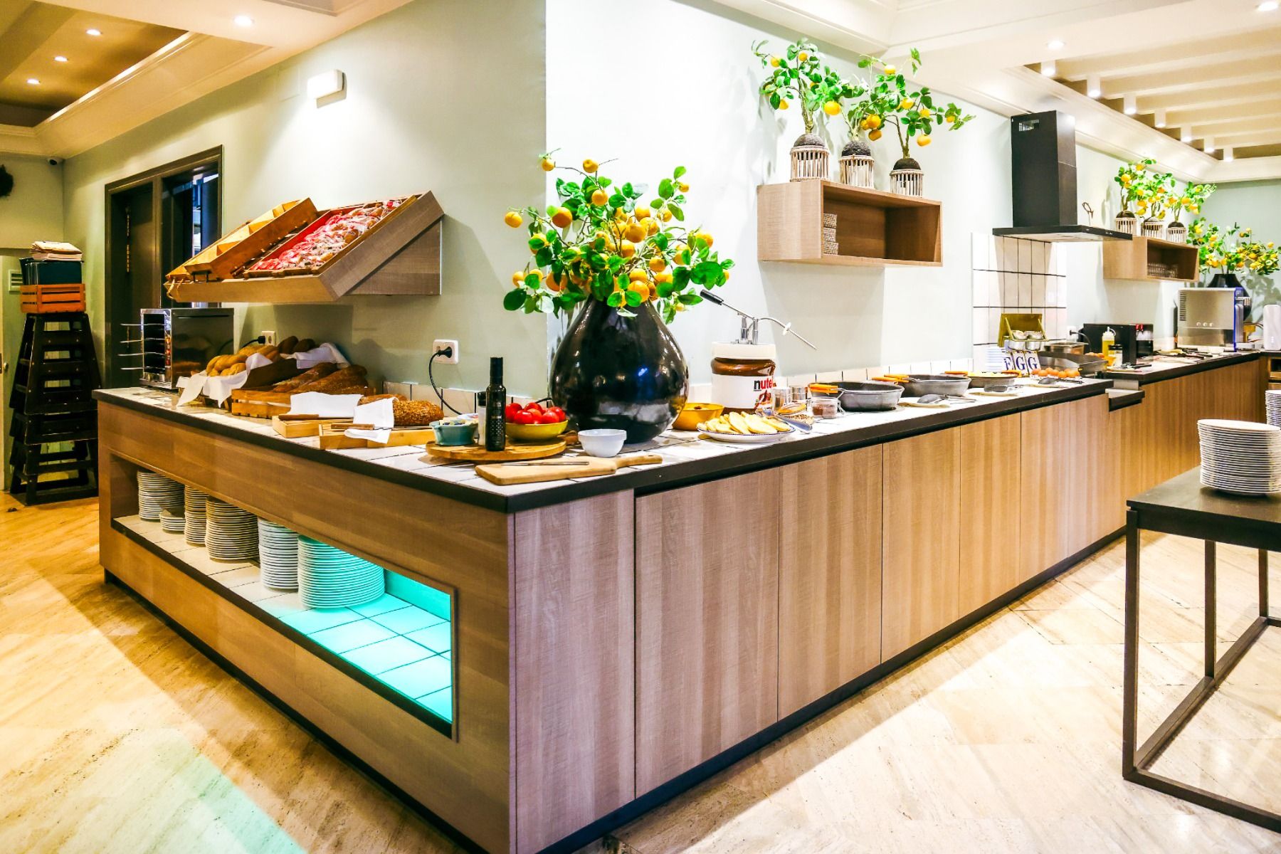 Ein modernes Buffet im Van der Valk Hotel BARCAROLA an der Costa Brava bietet hölzerne Theken mit Brot, Gebäck, Obst und Tellern. Grünpflanzen in dekorativen Vasen verleihen dem hellen, organisierten Raum Charme - ideal für einen Aufenthalt im Fußball-Tra