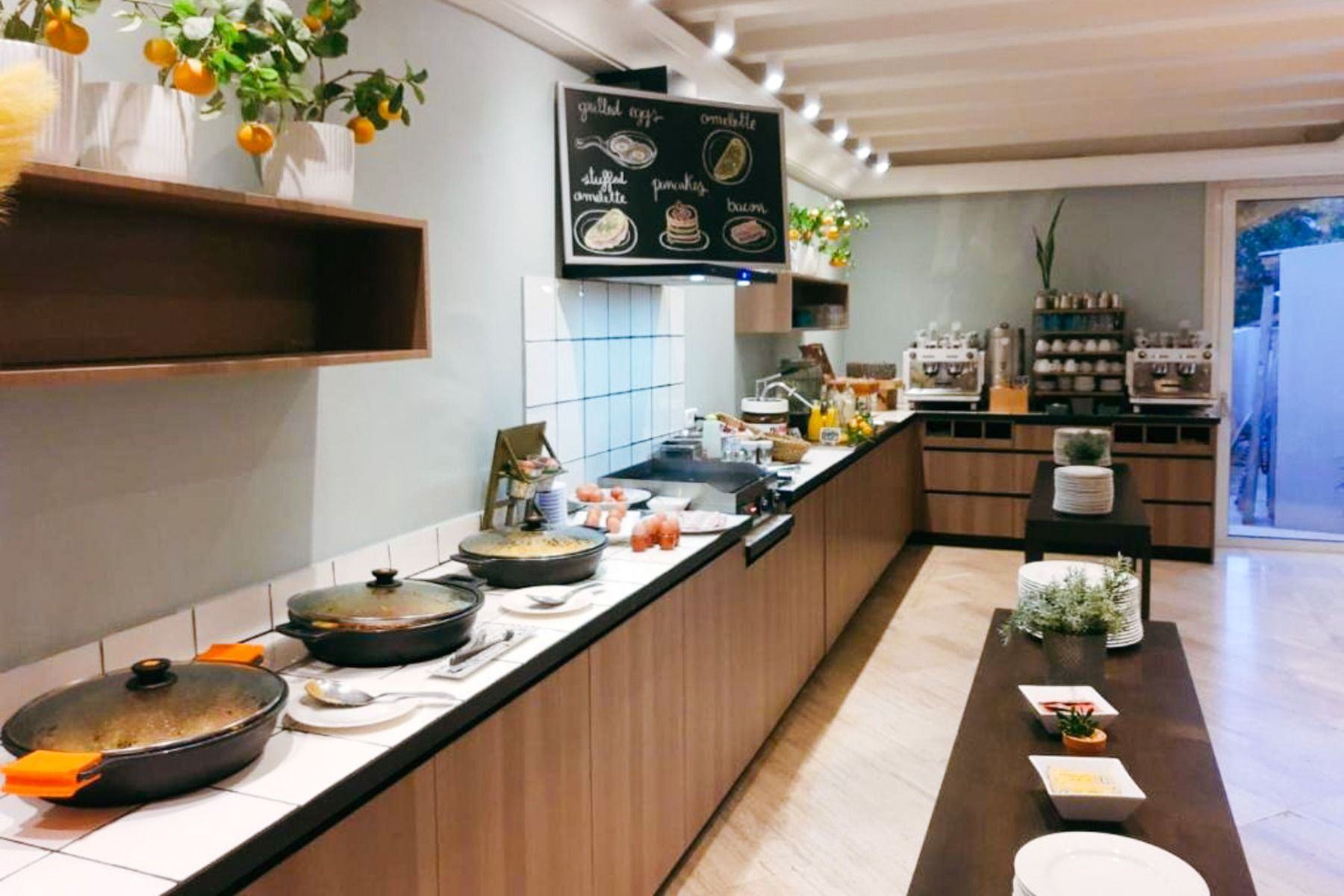 Erleben Sie im Van der Valk Hotel BARCAROLA eine moderne Küche mit Holzschränken, Kochtöpfen auf dem Herd, Topfpflanzen und Orangen - ideal für Gäste der Costa Brava oder für Mannschaften bei einem Fußball-Trainingslager.