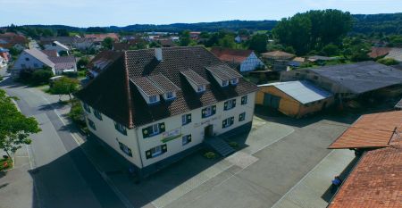 Ein gro&szlig;es, traditionelles Haus mit gr&uuml;nen Fensterl&auml;den und einem roten Ziegeldach steht an einer sonnigen Ecke, umgeben von Gr&uuml;n und einem eingez&auml;unten Garten. Auf einem Schild an der Veranda steht Gasthof Adler, der G&auml;sten gem&uuml;tliche Unterk&uuml;nfte bietet.