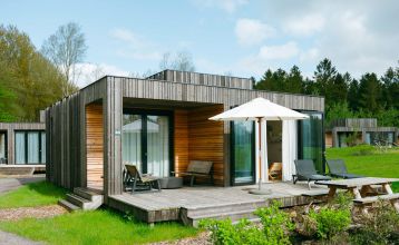 Im Resort De Parel öffnen sich moderne Holzkabinen mit großen Glastüren zu einem Deck mit Stühlen, einem Tisch und einem weißen Sonnenschirm - perfekt für Teambuilding oder Trainingslager im Grünen und ähnliche Kabinen unter teilweise bewölktem Himmel.