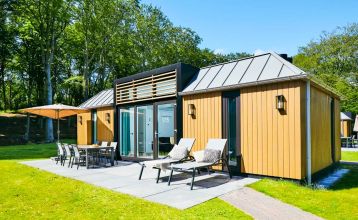 Modernes zweistöckiges Haus mit Metalldach, großen Fenstern und gelben Holzverkleidungen. Überdachte Terrasse mit Gartenmöbeln und Essbereich mit Blick auf einen grünen Rasen - perfekt zum Entspannen nach einem Tag in Duinrell.