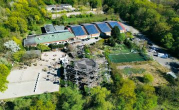 Luftaufnahme des von Bäumen umgebenen Sportparks Fun4You mit großem Solargebäude, Spielplatz, Teambuilding-Seilgarten, Sportplätzen, Parkplatz und bewaldeter Umgebung.