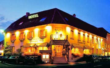 Der Hanauer Hof, ein großes traditionelles Hotel mit Ziegeldach und grünen Fensterläden, steht an einer Straßenecke in Appenweier. Autos sind draußen geparkt, Sonnenkollektoren säumen das Dach, und ein Kunstrasenplatz ist unter dem klaren blauen Himmel ni