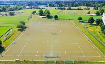 Luftaufnahme des Hotels Drei Rosen in Moosburg an der Isar – ein vierstöckiges, hellviolettes Hotel mit Sonnenkollektoren, Glasbalkonen und einem Außenbereich mit orangefarbenen Tischen und Stühlen, ideal für ein Fußballtrainingslager.