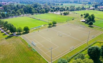 Luftaufnahme des Hotels Drei Rosen in Moosburg an der Isar – ein vierstöckiges, hellviolettes Hotel mit Sonnenkollektoren, Glasbalkonen und einem Außenbereich mit orangefarbenen Tischen und Stühlen, ideal für ein Fußballtrainingslager.
