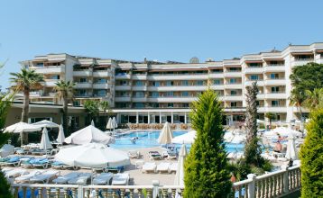 Das Linda Resort ist ein mehrstöckiges Hotel mit Blick auf einen Außenpool, der mit Liegestühlen, Sonnenschirmen und Palmen unter strahlend blauem Himmel ausgestattet ist.