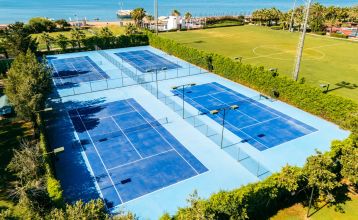 Luftaufnahme des Hotels Limak Arcadia mit Pools, Gärten, Sportplätzen und Strandkabinen - ideal für das Fußballtrainingslager Türkei - inmitten von Palmen und Bergen unter einem klaren Himmel.