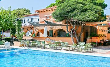 Das Van der Valk Hotel BARCAROLA ist ein 4-Sterne-Hotel im mediterranen Stil an der Costa Brava mit Balkonen mit Fahnen, einem klaren blauen Pool, Pooltischen mit Sonnenschirmen und Palmen unter einem strahlend blauen Himmel.