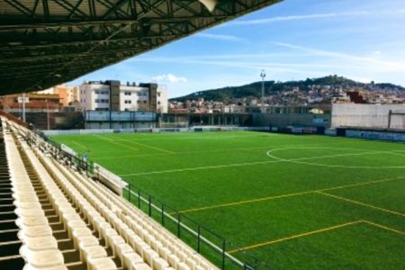 Die Reihen der leeren beigen Sitze im Campo Municipal de Fútbol Horta - Feliu i Codina überblicken ein grünes Kunstrasen-Fußballfeld, mit städtischen Gebäuden und einem baumbewachsenen Hügel im Hintergrund unter blauem Himmel.