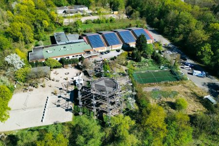 Luftaufnahme des von Bäumen umgebenen Sportparks Fun4You mit großem Solargebäude, Spielplatz, Teambuilding-Seilgarten, Sportplätzen, Parkplatz und bewaldeter Umgebung.