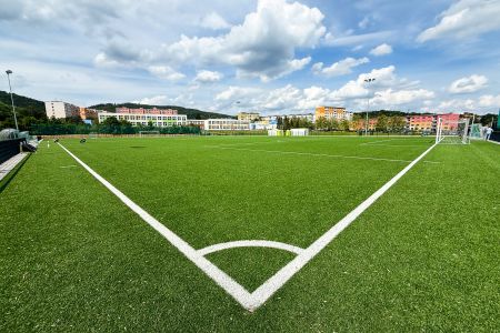 Der Kunstrasenplatz Klásterec besticht durch einen leuchtend grünen Kunstrasen-Fußballplatz mit strahlend weißen Begrenzungslinien, stabilen Torpfosten und farbenfrohen Gebäuden im Hintergrund unter einem teilweise bewölkten Himmel.