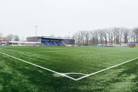 SV Grol Kunstrasen 1 hat einen grünen Kunstrasenplatz mit weißen Begrenzungslinien und eine überdachte Tribüne mit roten und blauen Sitzen. Im Hintergrund sind Bäume und Gebäude bei bedecktem Himmel zu sehen.