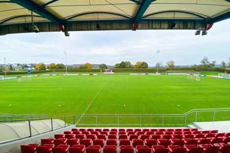 Blick von der Tribüne des leeren Fußballplatzes des SV Güssing mit grünem Rasen, kleinen Toren, roten Stadionsitzen unter einem überdachten Dach an einem bewölkten Tag; im Hintergrund sind Bäume und Häuser zu sehen.
