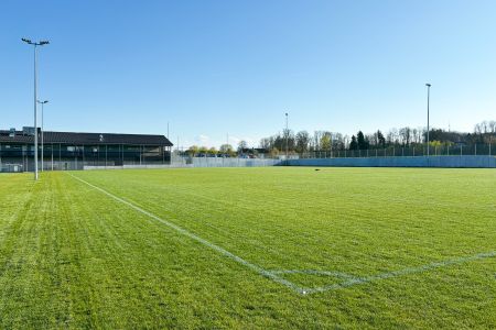 Luftaufnahme des Schechener Hauptplatzes, ein grüner Fußballplatz neben einem Gebäude und einem Parkplatz, umgeben von Straßen, Feldern und einem Dorf mit Bergen in der Ferne unter einem klaren Himmel.