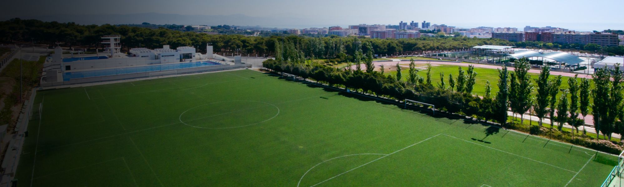 Kunstrasen-Fußballplatz auf Sardinien mit markierten Spielfeldlinien und Umzäunung, im Hintergrund Berge und Gebäude.