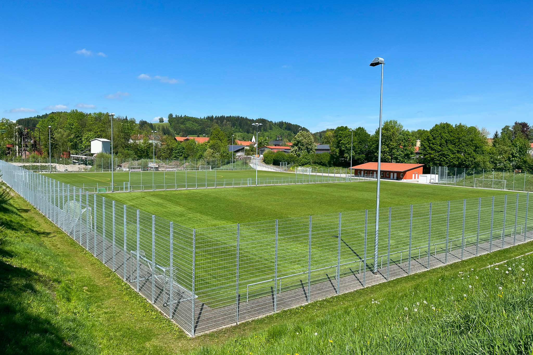 Fußballtrainingsplatz auf Sardinien mit grünem Kunstrasen, Tor, Bänken und Flutlichtmasten, umgeben von Netzen und Berglandschaft.