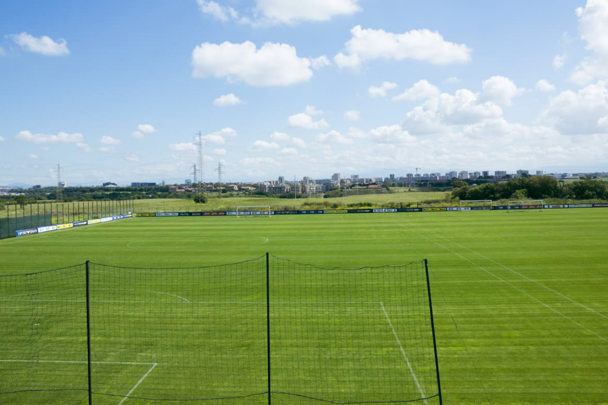 Fußballtrainingsplatz auf Sardinien mit grünem Kunstrasen, Tor, Bänken und Flutlichtmasten, umgeben von Netzen und Berglandschaft.