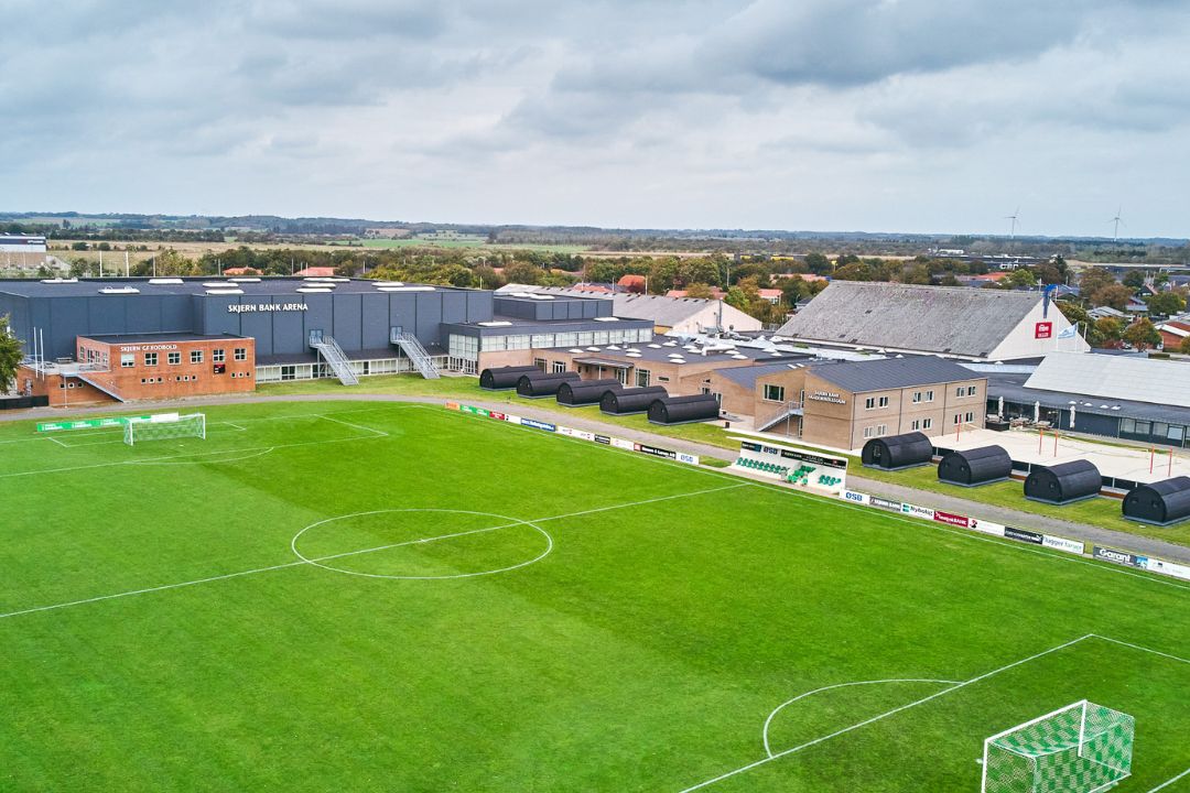 Fußballplatz mit angrenzender Sporthalle und modernen Unterkunftsmodulen in Süddänemark, umgeben von ländlicher Landschaft und kleinen Gebäuden.