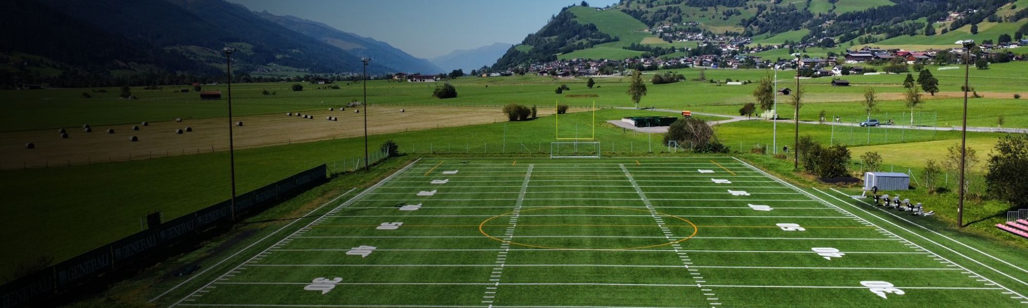 Kunstrasen-Fußballplatz auf Sardinien mit markierten Spielfeldlinien und Umzäunung, im Hintergrund Berge und Gebäude.