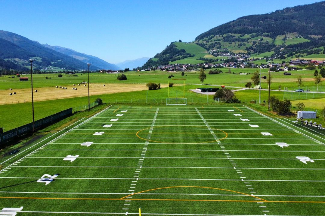 Fußballtrainingsplatz im Zillertal mit grünem Kunstrasen, Tor und Flutlichtmasten, umgeben von blauem Himmel und Berglandschaft.