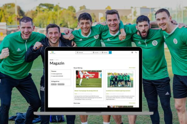 Five men in matching green tracksuits are standing outside on a field, laughing and celebrating, behind a large computer monitor displaying a German-language sports website.