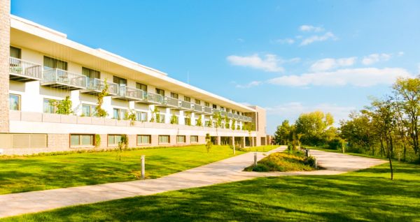 The Akademia Sporthotel is a modern hotel with balconies, located right next to a paved walkway and a lush green lawn. It is surrounded by trees and sits beneath a bright blue sky dotted with scattered clouds. Here, guests can enjoy a relaxing spa area.