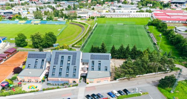 Aerial view of Hotel Benica*** featuring two artificial turf pitches, buildings with solar panels, a parking lot with cars, and surrounding trees and urban area—perfect for a training camp or wellness area.