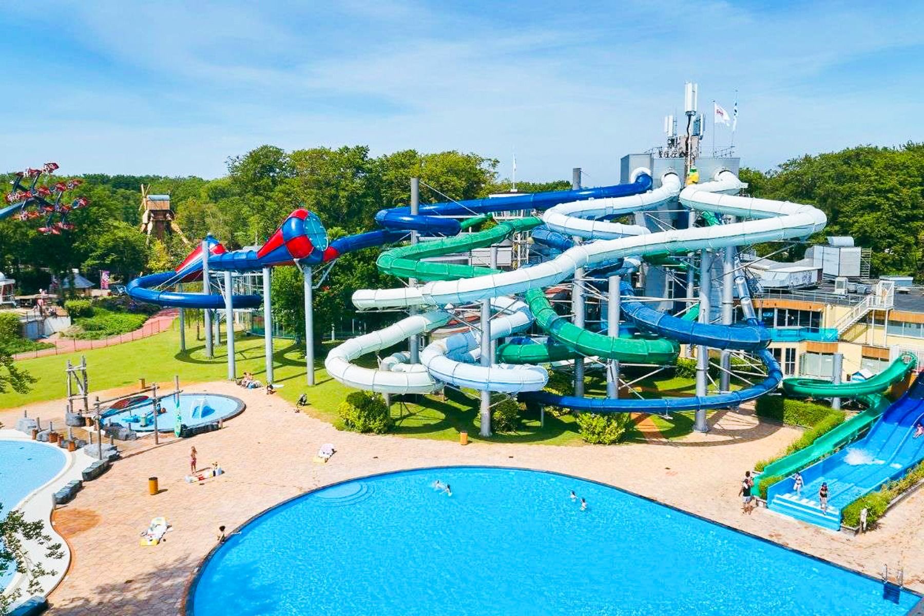 Duinrell boasts a large outdoor water park with intertwining slides, blue pools, and guests swimming or relaxing. Surrounded by green trees and under a sunny sky, it showcases impressive sports facilities in a scenic setting.