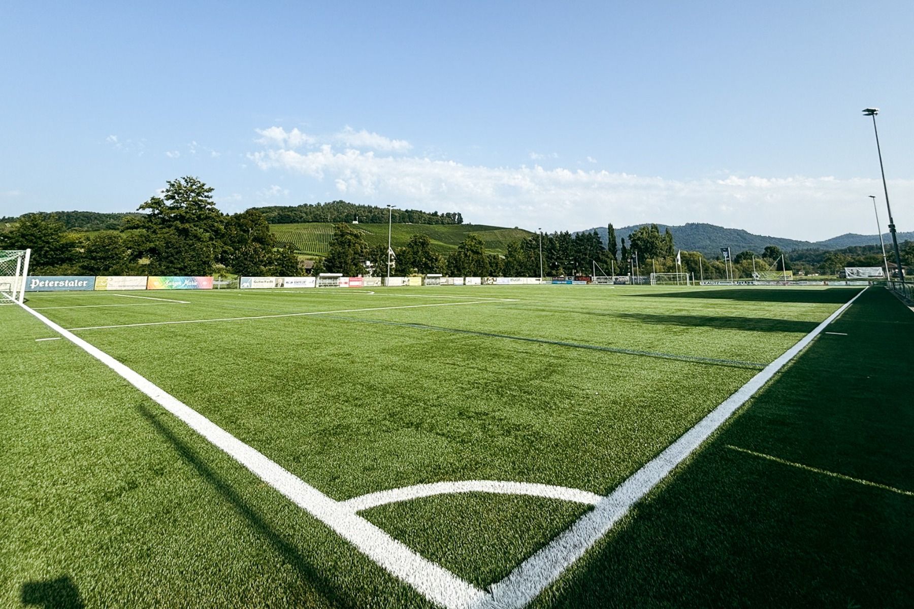 A wide-angle view of an artificial turf pitch at Hanauer Hof with white boundary lines, surrounded by trees and hills under a clear blue sky. Advertising boards line the fence, making it ideal for a training camp setting.