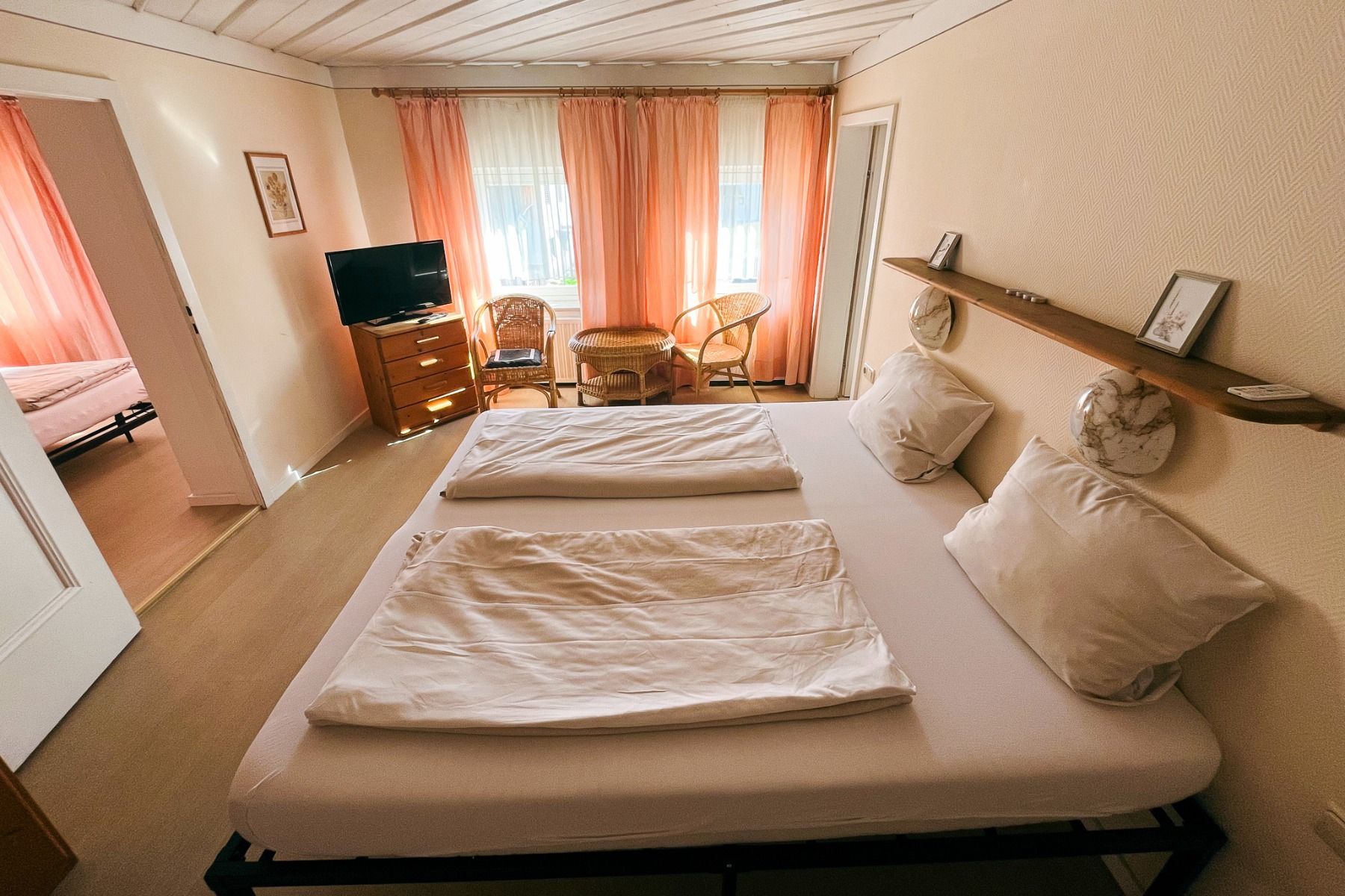 Egger Stüberl: A cozy bedroom, ideal for training camps, with two single beds with white bed linen, a TV on a wooden chest of drawers, wicker armchairs by the window with peach-colored curtains, and an adjoining room with a visible extra bed.