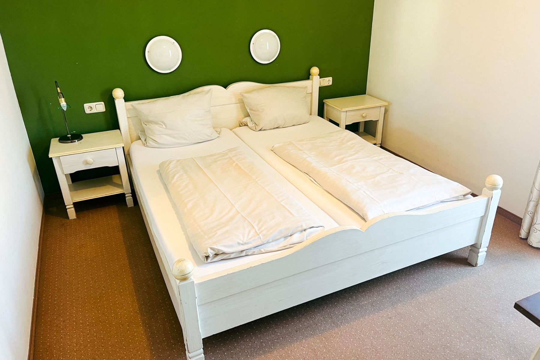 The Egger Stüberl has a neatly made white double bed with pillows and comforters in front of a green wall, flanked by white bedside tables with lamps and decorative signs above - perfect for relaxing after a day at training camp.