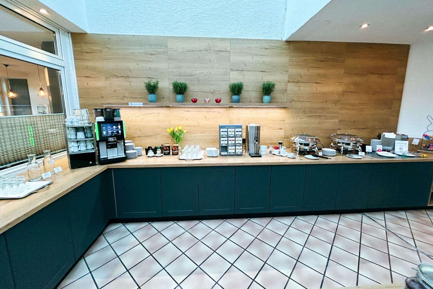 Hotel Empfinger Hofs frokostbuffé består av en kaffemaskin, te-stasjon, glass, tallerkener og tildekkede matbrett på en disk ved en trevegg, supplert av potteplanter og rød dekor over.