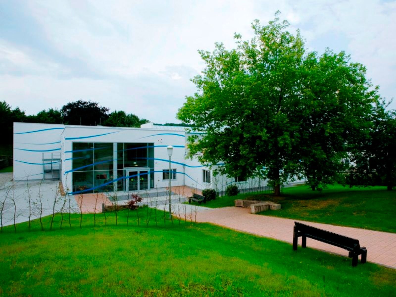 The sports and seminar center is a modern white building with blue accents, perfect for training. It stands behind a green lawn and a tree, with a black bench on the paved path in front of it and a cloudy sky above us.