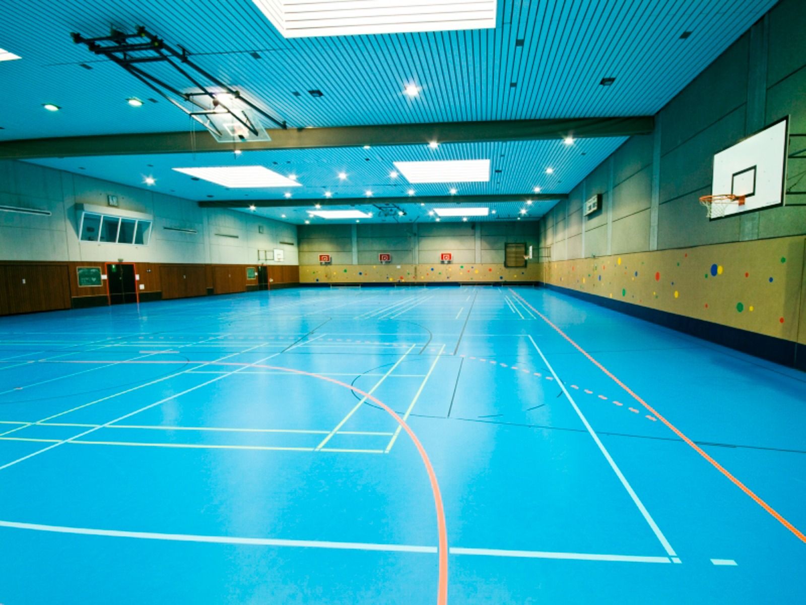 The Sport & Seminar Center features a spacious indoor gymnasium with a bright blue floor, basketball hoops, and marked sports lines—perfect for training camps or fitness activities. Skylights illuminate its light brown and gray paneled walls.