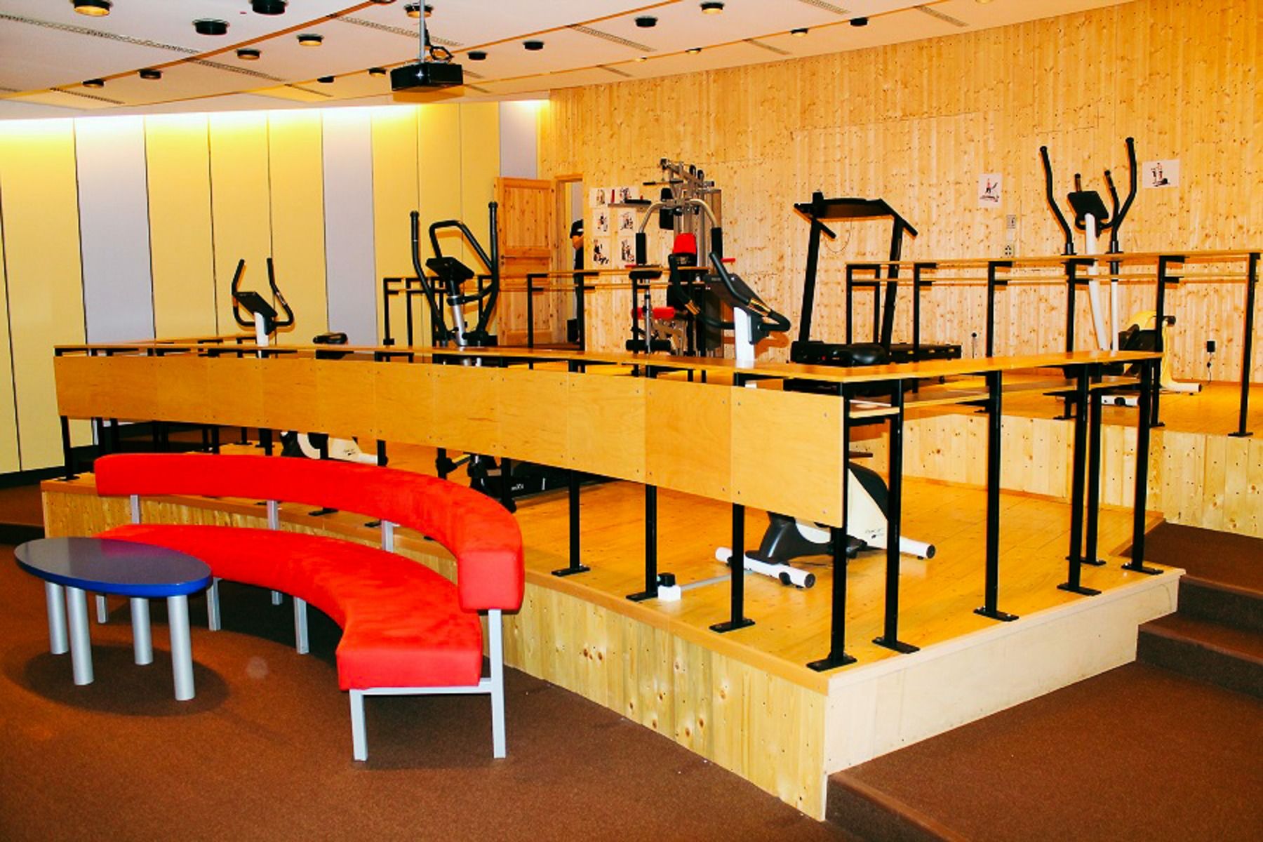 The Sport & Seminar Center features a modern exercise room with elliptical machines and treadmills on a raised wooden platform, a curved red bench with small tables in the foreground, and light wood-paneled walls.
