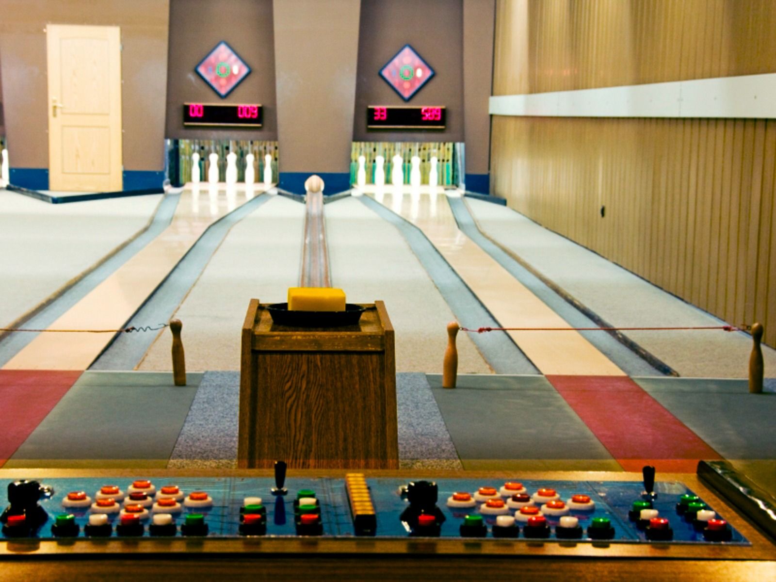 A view of two bowling lanes with pins set up, seen from behind a wooden control panel at the Sport & Seminar Center—an inviting, well-lit area often found in fitness and wellness facilities, currently empty.