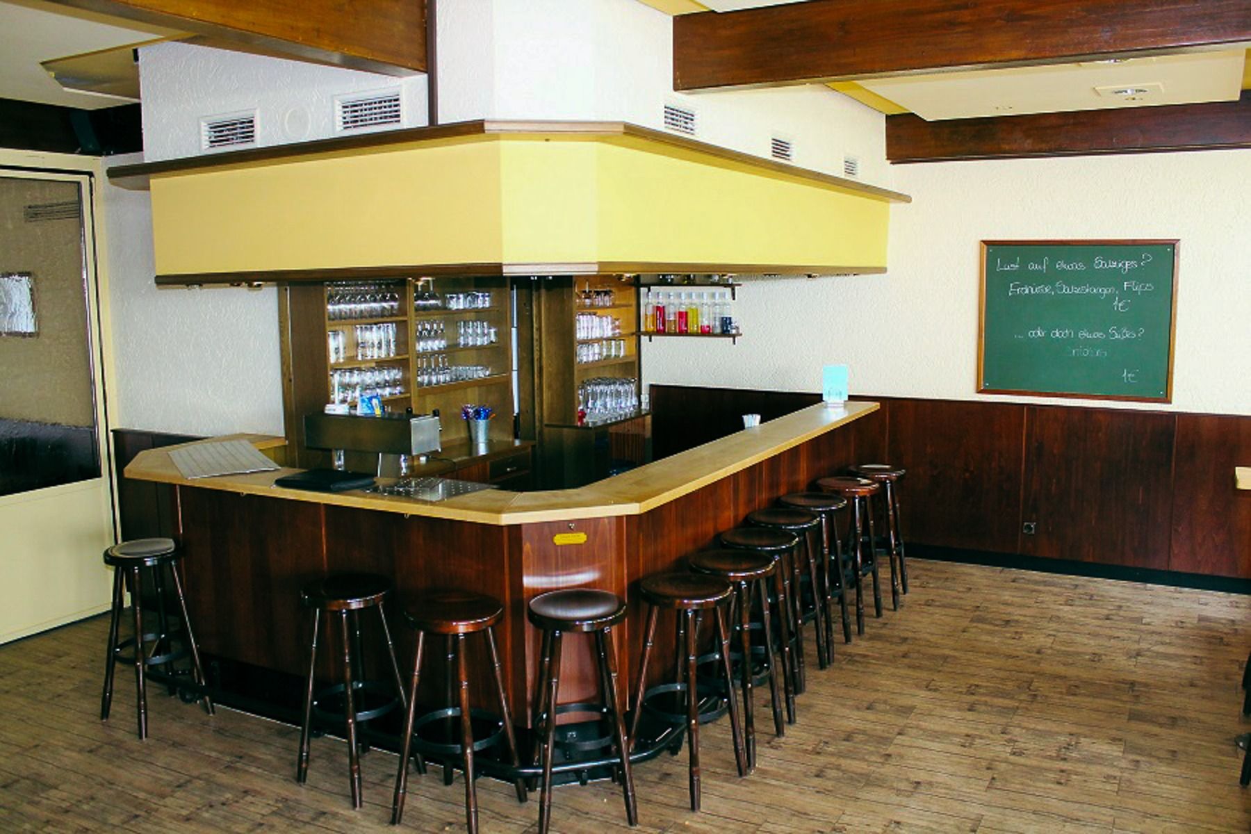 A cozy, empty bar with wooden counter and bar stools, glasses hanging above and bottle racks behind - perfect as a relaxation area in the fitness and wellness area of the Sport & Seminar Center or as part of your training camp destination.