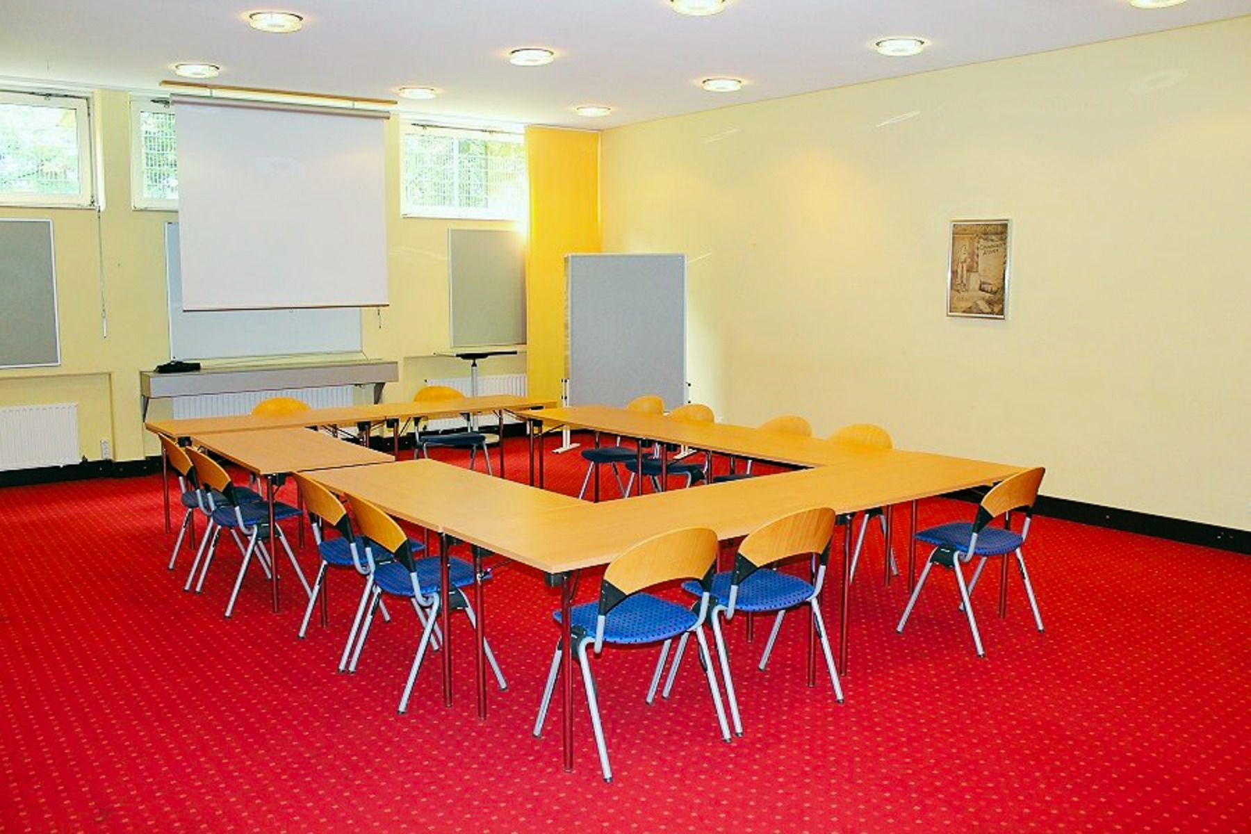 The Sport & Seminar Center features a bright conference room with U-shaped tables, blue-seated wooden chairs, projector screen, red carpet, light yellow walls, and high windows—perfect for training camps or wellness workshops.