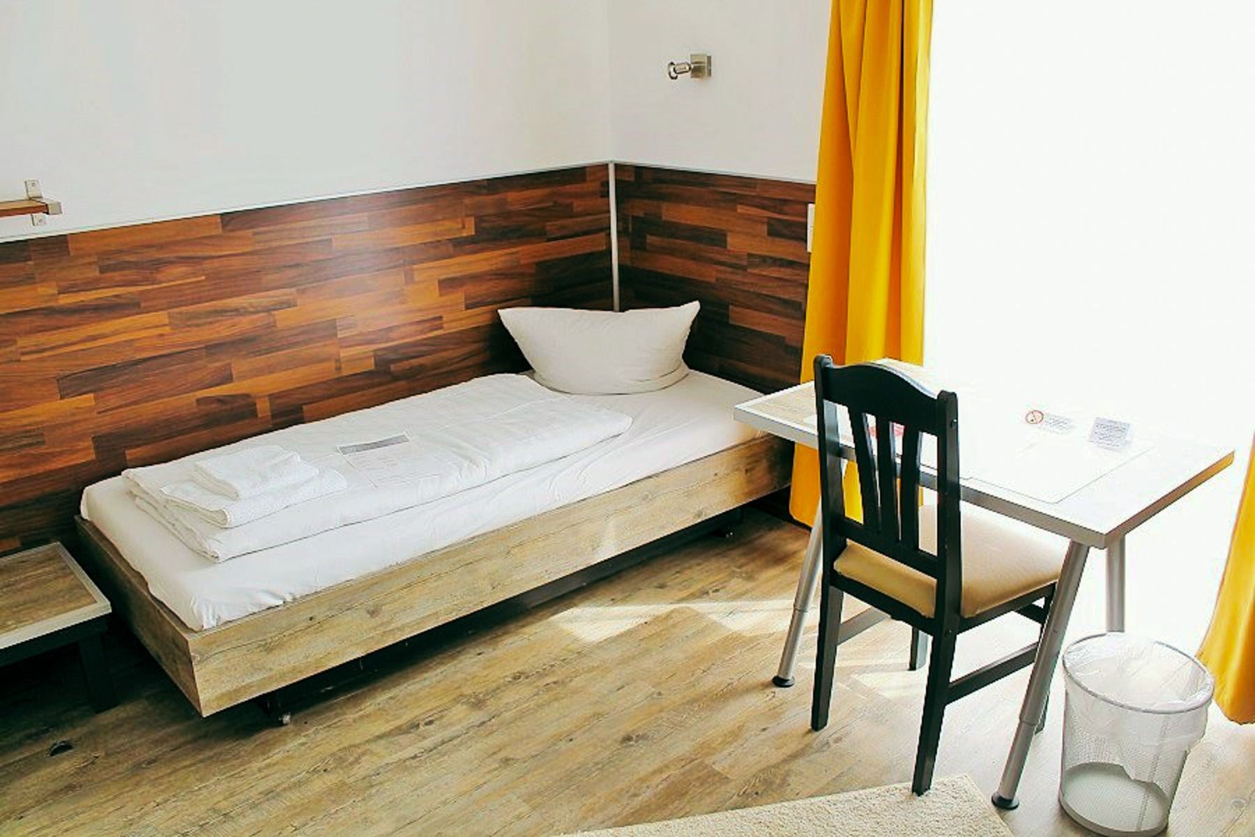 A cozy single room in the sports and seminar center features white bedding, wooden accents, a desk and chair, a wastebasket and yellow curtains framing a large, sunlit window - perfect for relaxing after training camp.