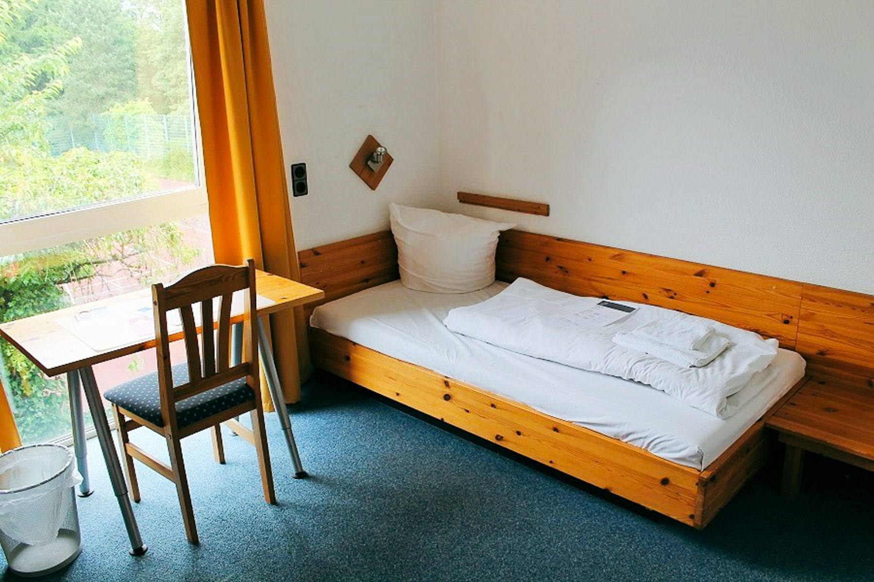 A small, tidy room in the sports and seminar center with a single wooden bed, white bedding, desk and chair by a window with yellow curtains and easy access to the conference rooms and the fitness and wellness area on the blue carpeted floor.