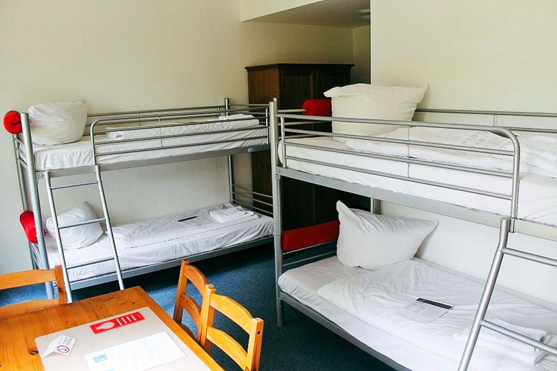 A bright room in the Sports & Seminar Center with two metal bunk beds with white bedding, a wooden table and chairs in the foreground and papers and a red object on the table gives the impression of a training camp.