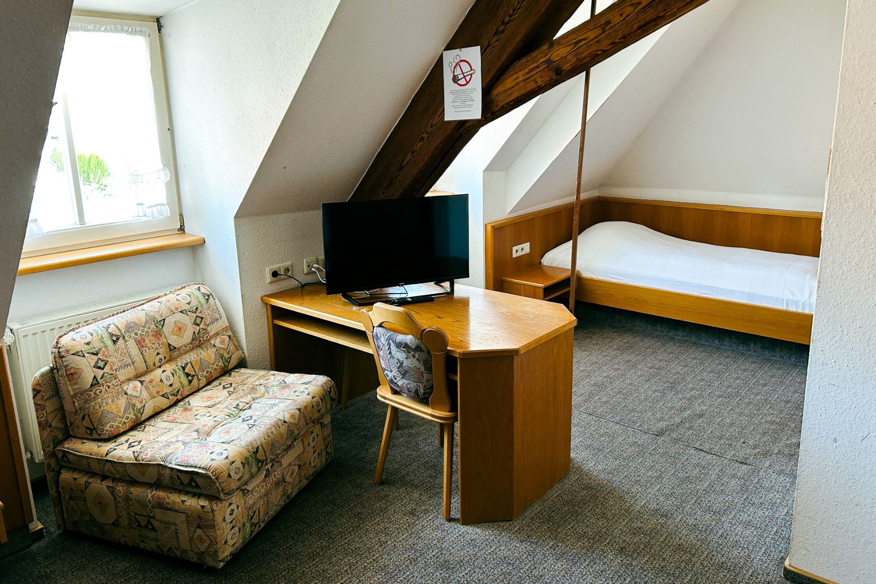 Hotel Adler’s small attic room with slanted ceilings features a single bed, wooden desk with TV and chair, patterned armchair, and a window for natural light—providing cozy accommodation.