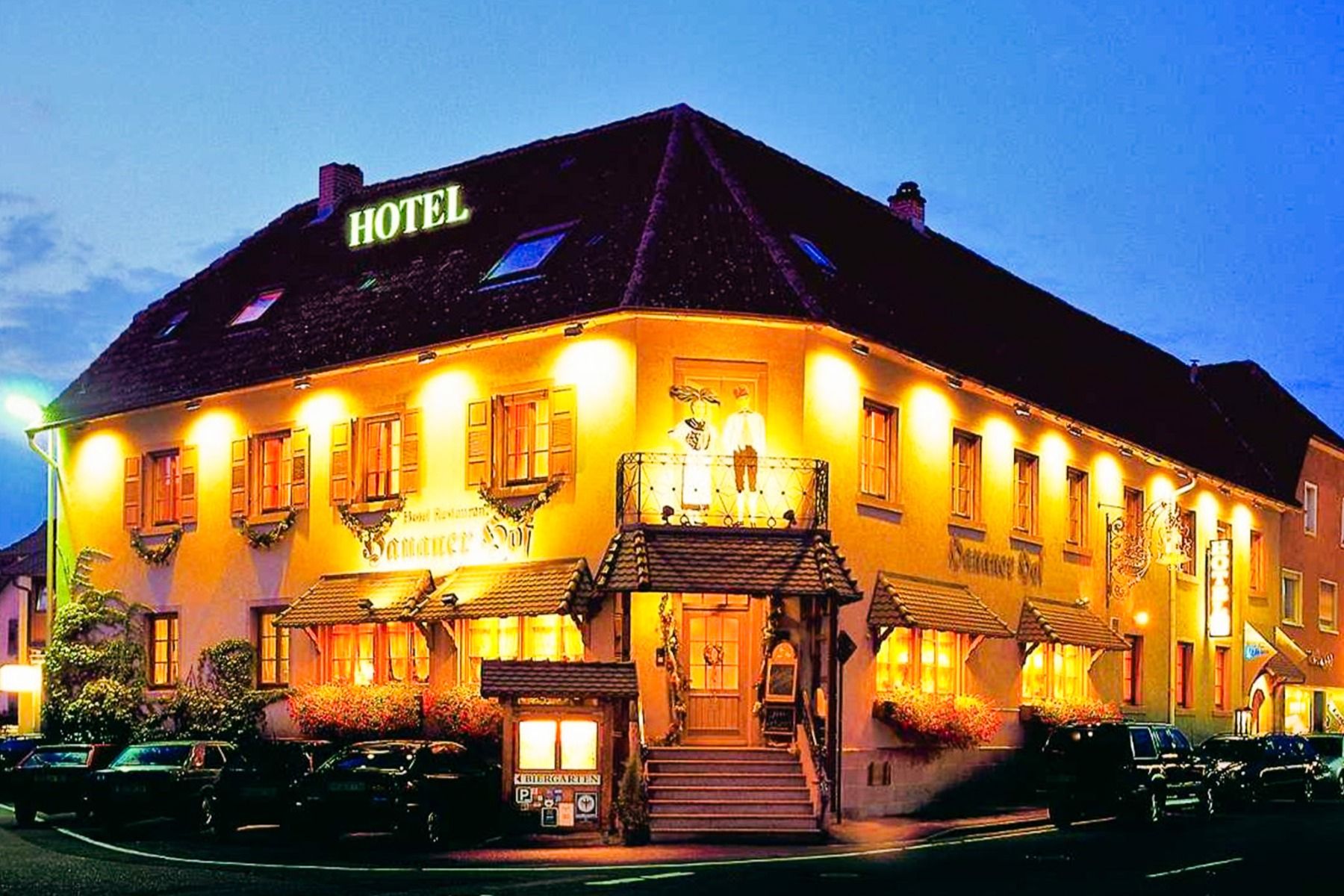 Hanauer Hof: A warmly lit, yellow hotel with flower boxes and a glowing HOTEL sign on the roof, photographed at dusk—an ideal choice for a training camp or stay near the artificial turf pitch in Appenweier.