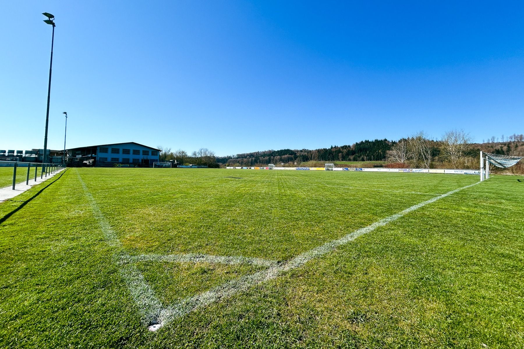 Hotel Adler overlooks an empty outdoor soccer field with a neatly mowed green pitch, white corner lines, and goalposts—an ideal family spot surrounded by trees, hills, and a scenic building in the background.